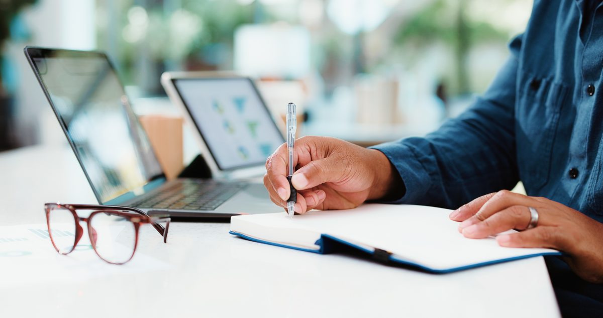 Person writing in a notebook beside a laptop and tablet