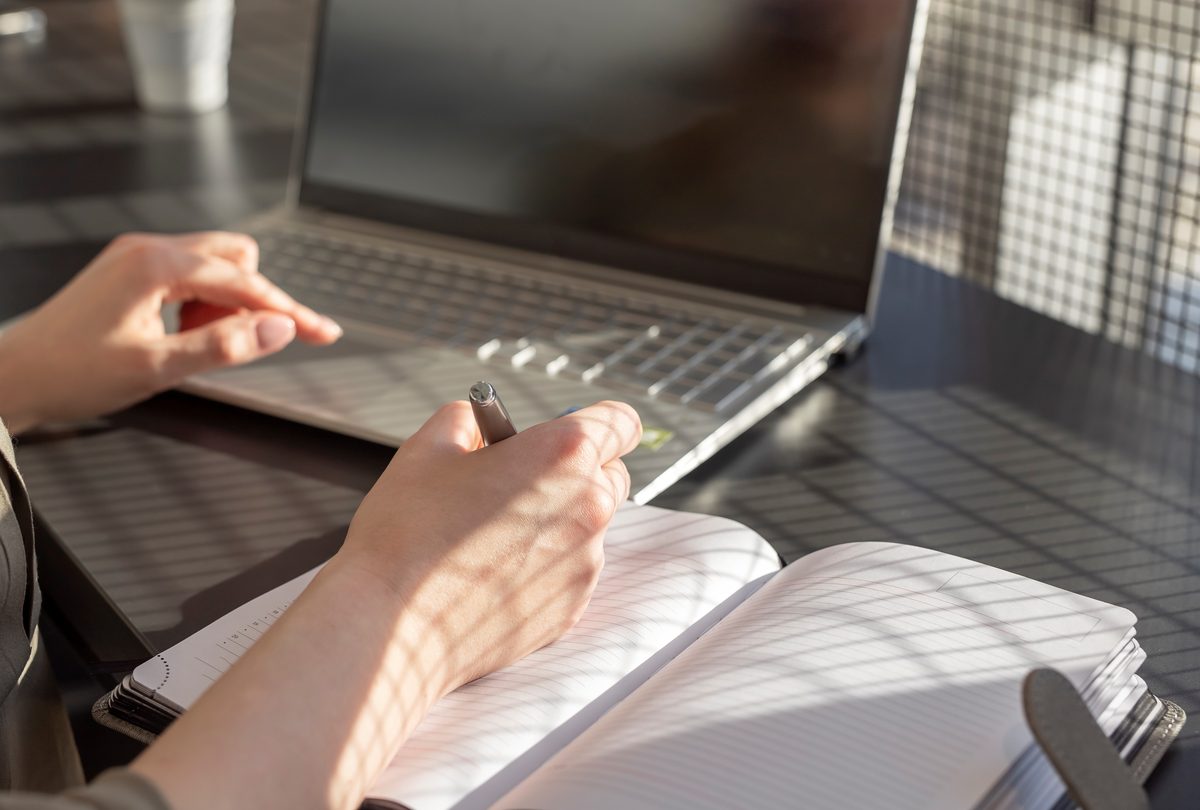 Hands writing notes in a notebook beside a laptop